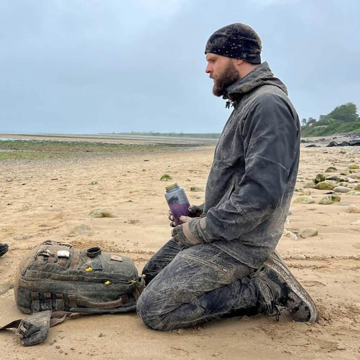 Man kneeling on sandy, rocky beach with muddy GORUCK backpack and water bottle, outdoor gear