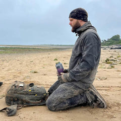 Man kneeling on sandy, rocky beach with muddy GORUCK backpack and water bottle, outdoor gear