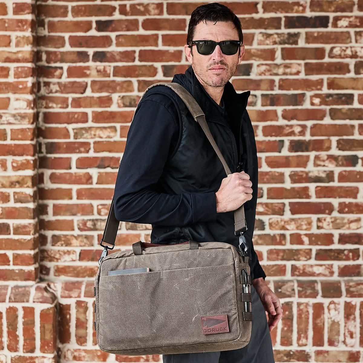 Man wearing sunglasses carrying a GORUCK messenger bag in front of a brick wall