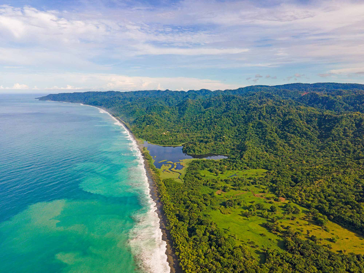 Aerial view of a lush green coastline with turquoise ocean waves and dense forest under a partly cloudy sky.