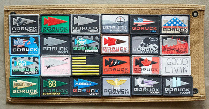 A display panel with 24 colorful GORUCK event patches arranged in a neat grid on a tan background.
