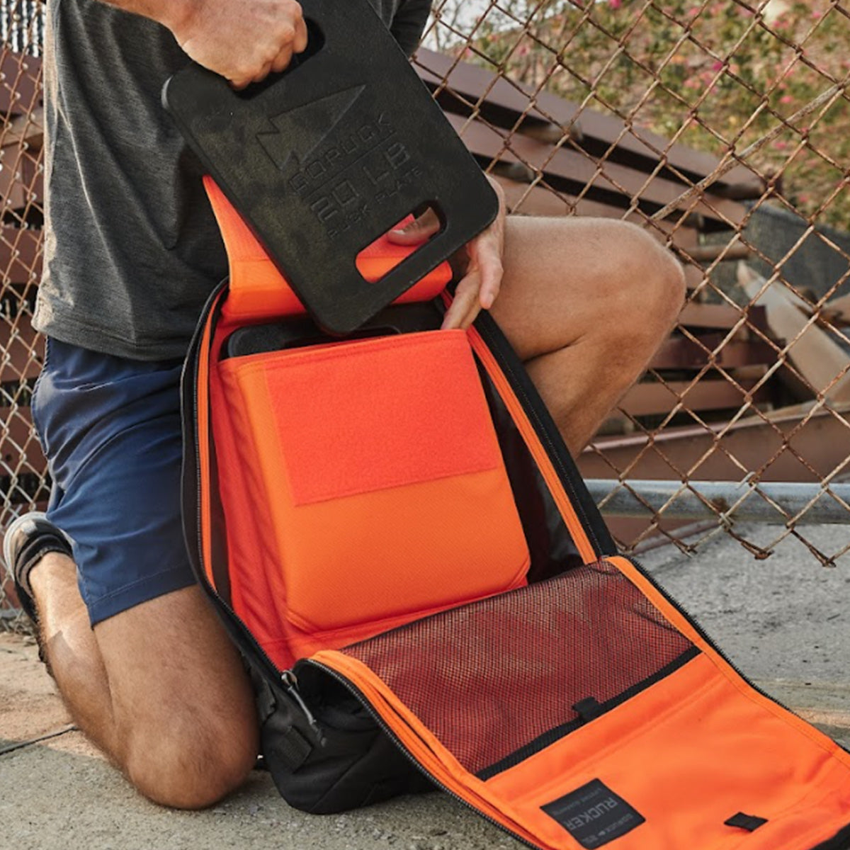 A person places a 20 lb weight plate into an orange backpack near a chain-link fence.