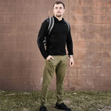 Man with a backpack stands on grass in front of a brick wall, wearing a black top and Men’s Training Performance Joggers in olive, made in the USA.