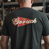 Man wearing the GORUCK Bud Tee - Tri-BlendX, featuring a bold red and gold "Goruck" logo on the back. Ideal for everyday wear and comfort indoors.