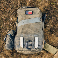 A muddy Rucker 5.0 rucksack rests on dirt, featuring a water bottle attachment and top patch—designed for rucking adventures with built-in durable lumbar support.