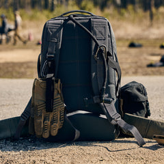 The Rucker 5.0, a black tactical rucksack with gloves and gear attached, sits outdoors on the ground, ready for any adventure and designed to provide reliable lumbar support.
