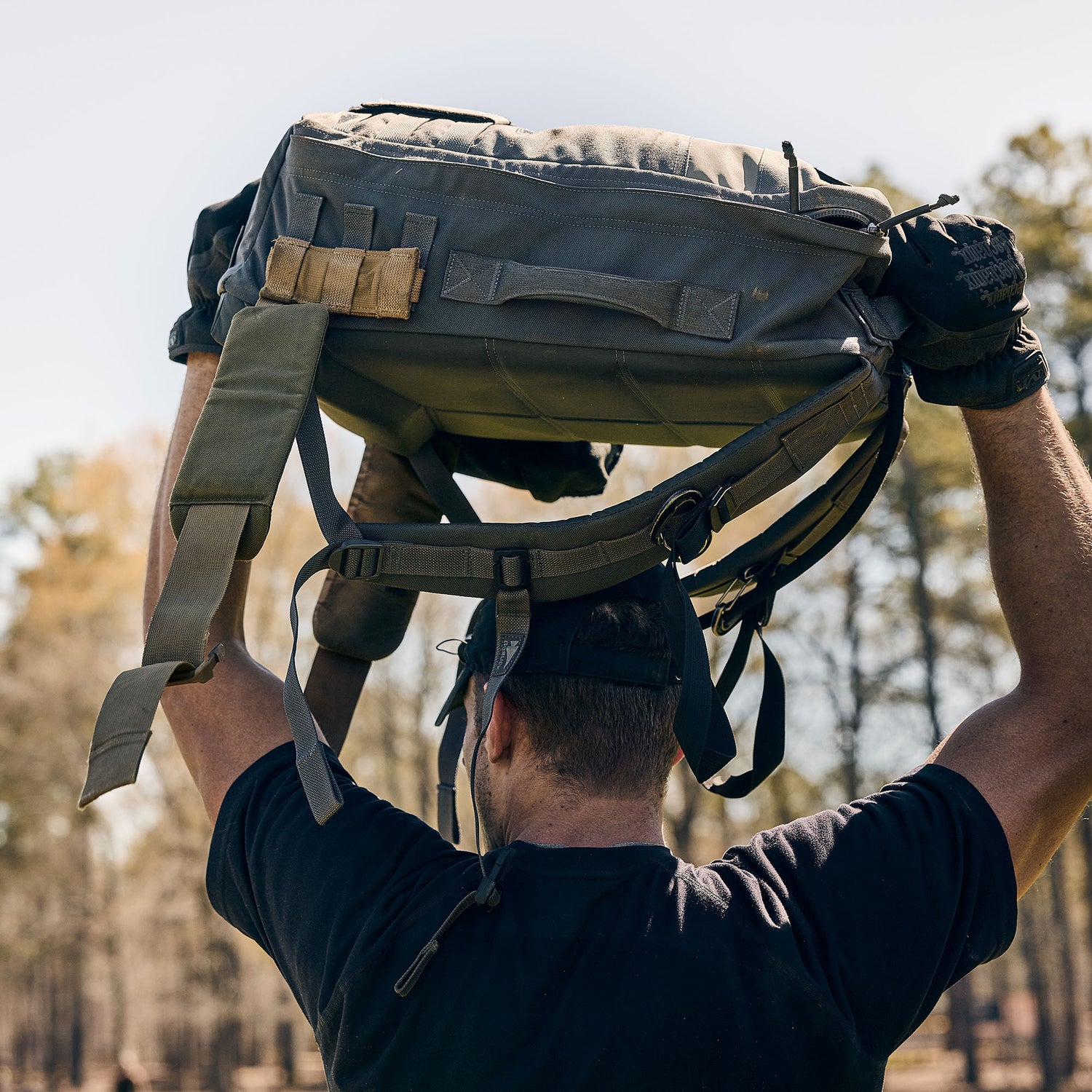 Wearing a black shirt and gloves, a person lifts the Rucker 5.0 overhead outdoors with trees behind them, highlighting their strength and the advantages of rucking.