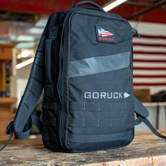 A black Rucker 5.0 rucksack with an American flag patch sits on a wooden table indoors, ready for your next adventure and designed with excellent lumbar support.