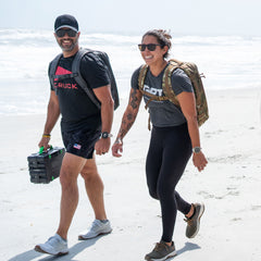 Two people with rucksacks walk and smile on a sunny beach, enjoying the ocean breeze and the superior lumbar support of the Rucker 5.0.