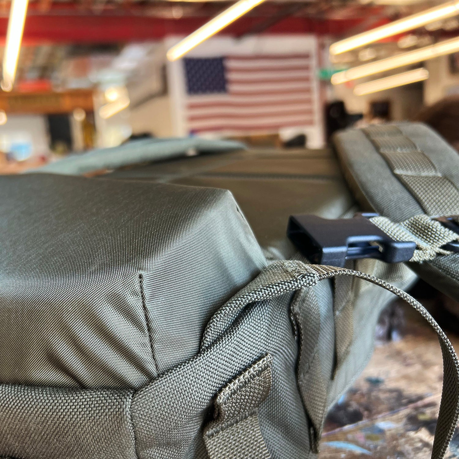 Close-up of the olive green Rucker 5.0 rucksack with lumbar support, with an American flag hanging in the blurred background.