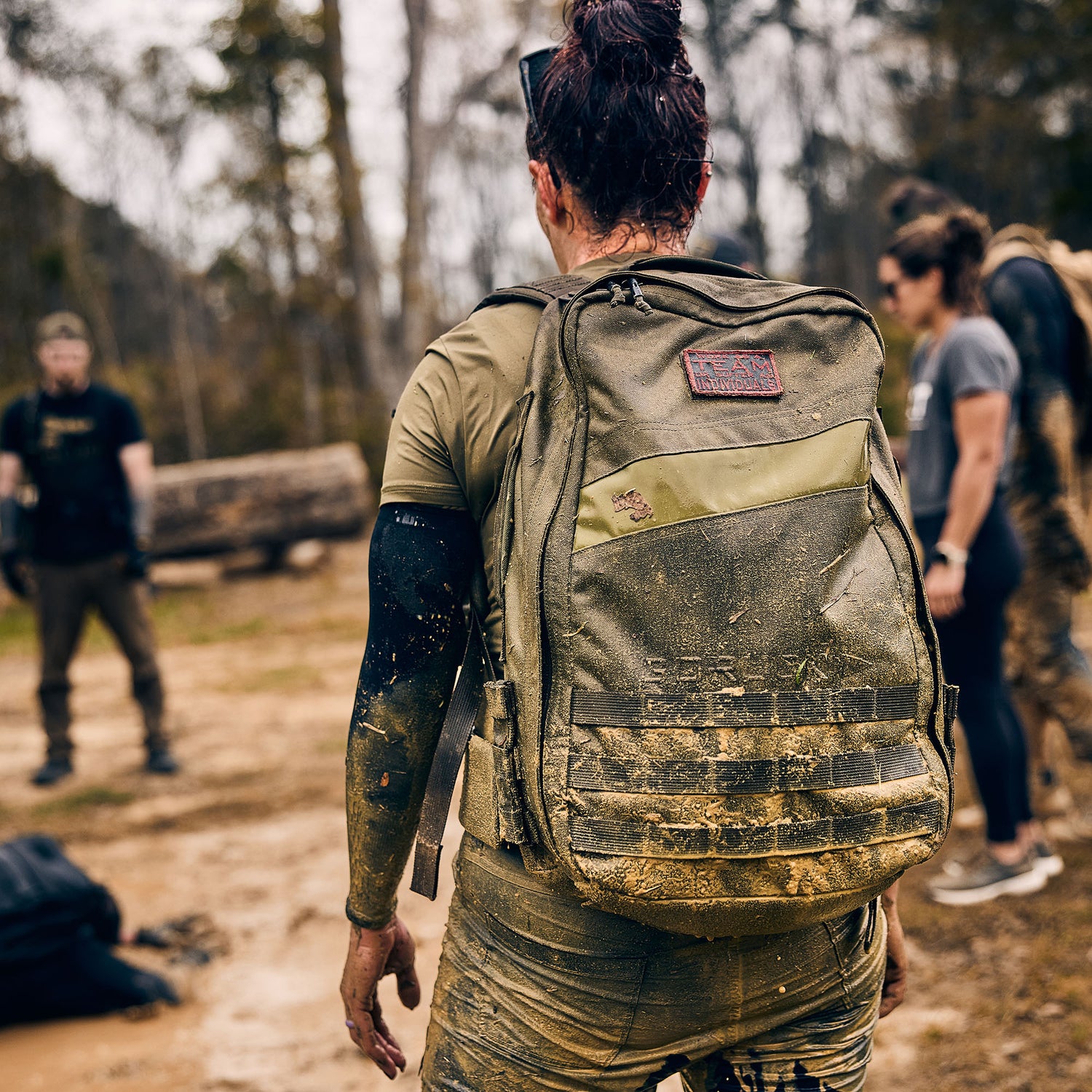 Wearing the Rucker 5.0, a person with a muddy rucksack stands outside among others in athletic clothing on a dirt path, ready for a day of rucking.