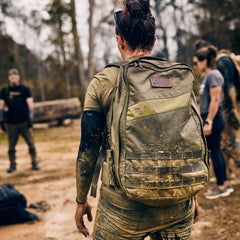 Wearing the Rucker 5.0, a person with a muddy rucksack stands outside among others in athletic clothing on a dirt path, ready for a day of rucking.