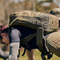 Wearing the Rucker 5.0 with lumbar support, a person in outdoor gear performs push-ups on grass during a fitness rucking session.
