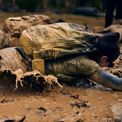 Wearing the Rucker 5.0, a person crawls through thick mud with a large rucksack, splashing mud during an intense outdoor rucking challenge.