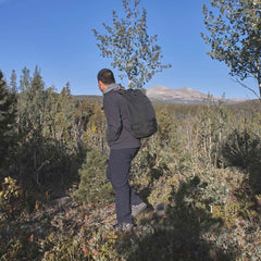 Man rucking in GORUCK gear and pants on a mountain trail surrounded by trees and blue sky.
