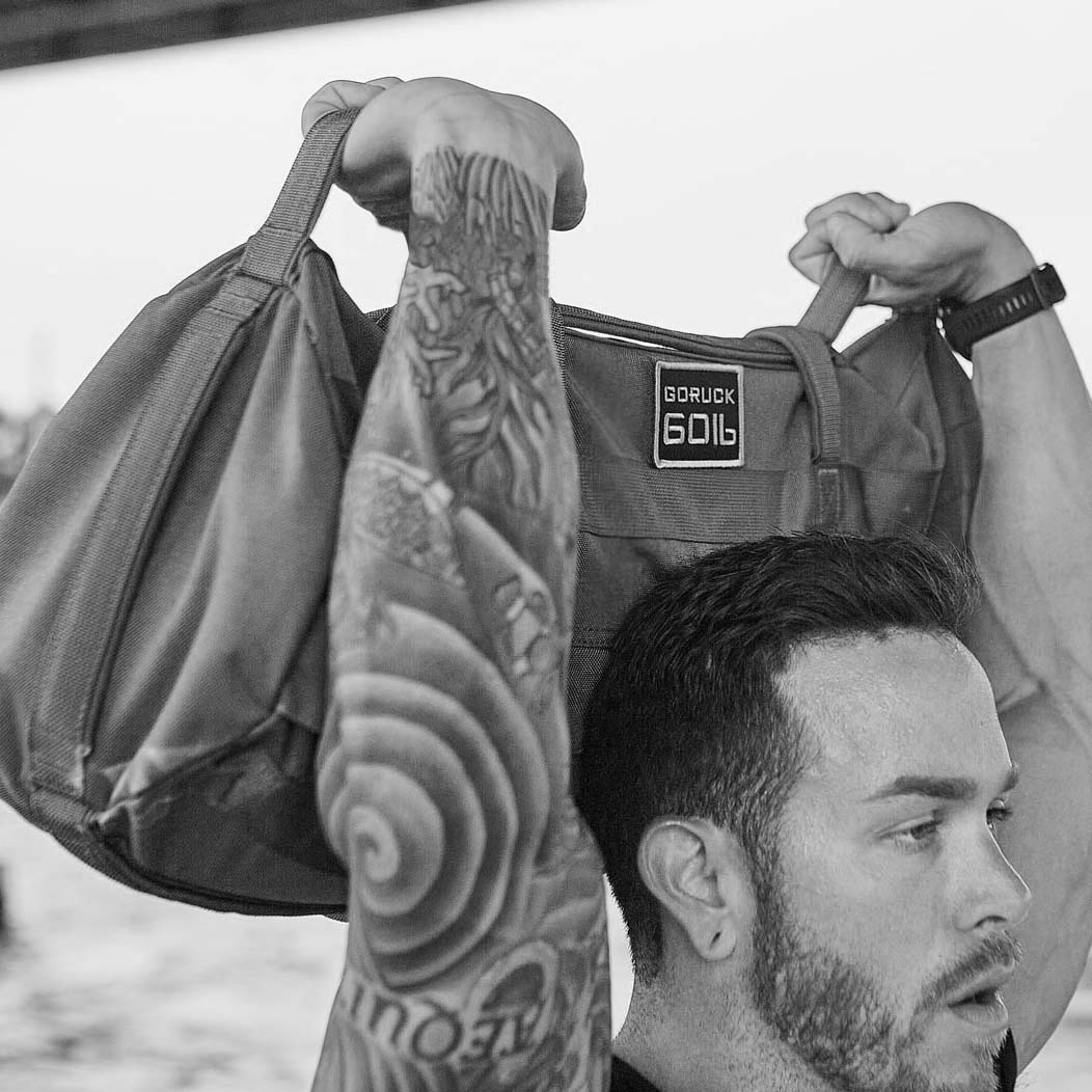 Man lifting a GORUCK 60LB sandbag overhead, showing a tattooed arm, in black and white.