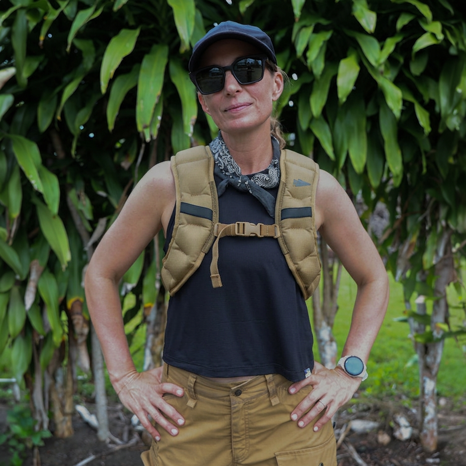 Woman outdoors sporting sunglasses, a cap, bandana, black tank top, and brown cargo pants—ready for adventure with her Spy Ruck | Women's Weighted Vest.