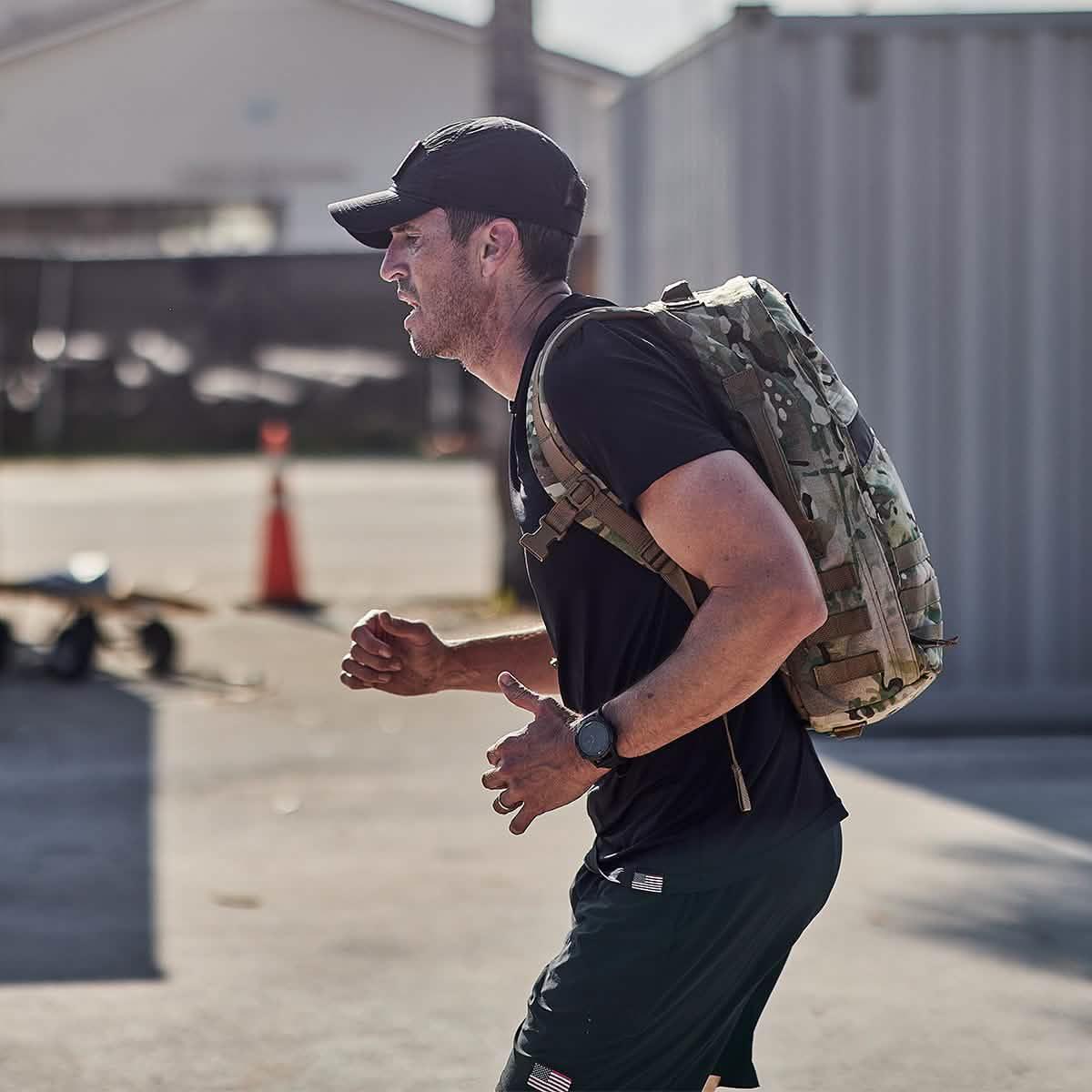 Man rucking outdoors with GORUCK camo backpack, wearing athletic gear and a cap