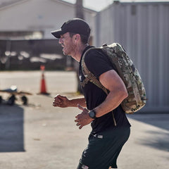 Man rucking outdoors with GORUCK camo backpack, wearing athletic gear and a cap
