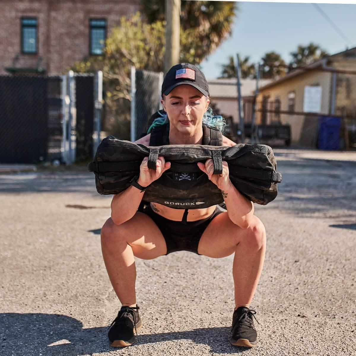 Woman doing outdoor squat with GORUCK sandbag, wearing athletic gear and a cap