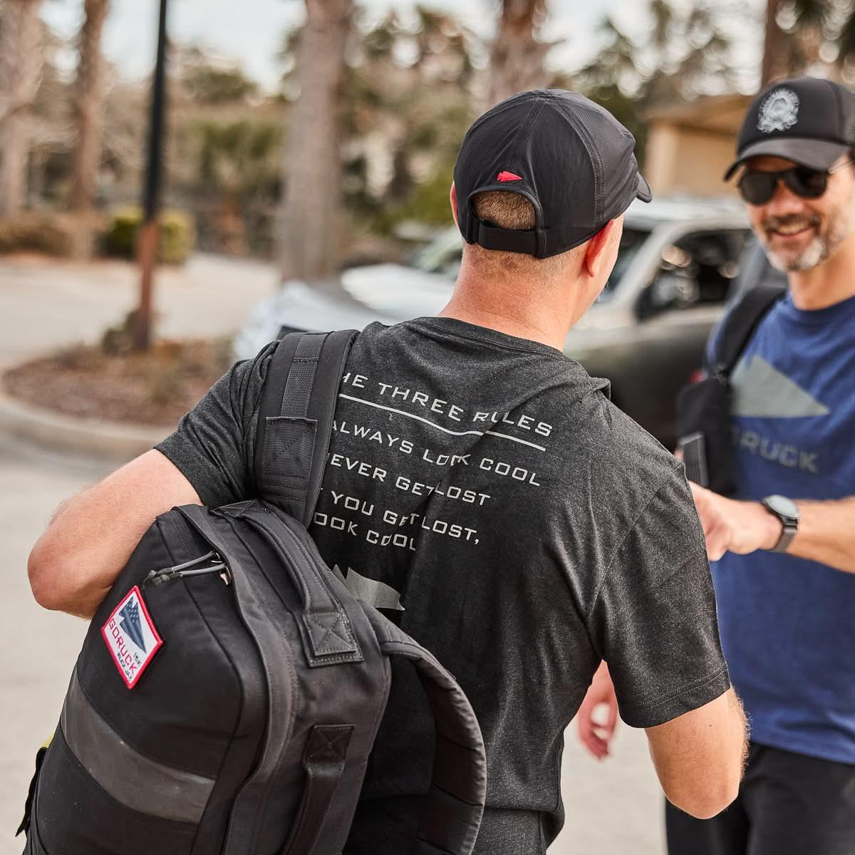 Man in GORUCK The Three Rules tee carrying black ruck, talking outdoors with another man