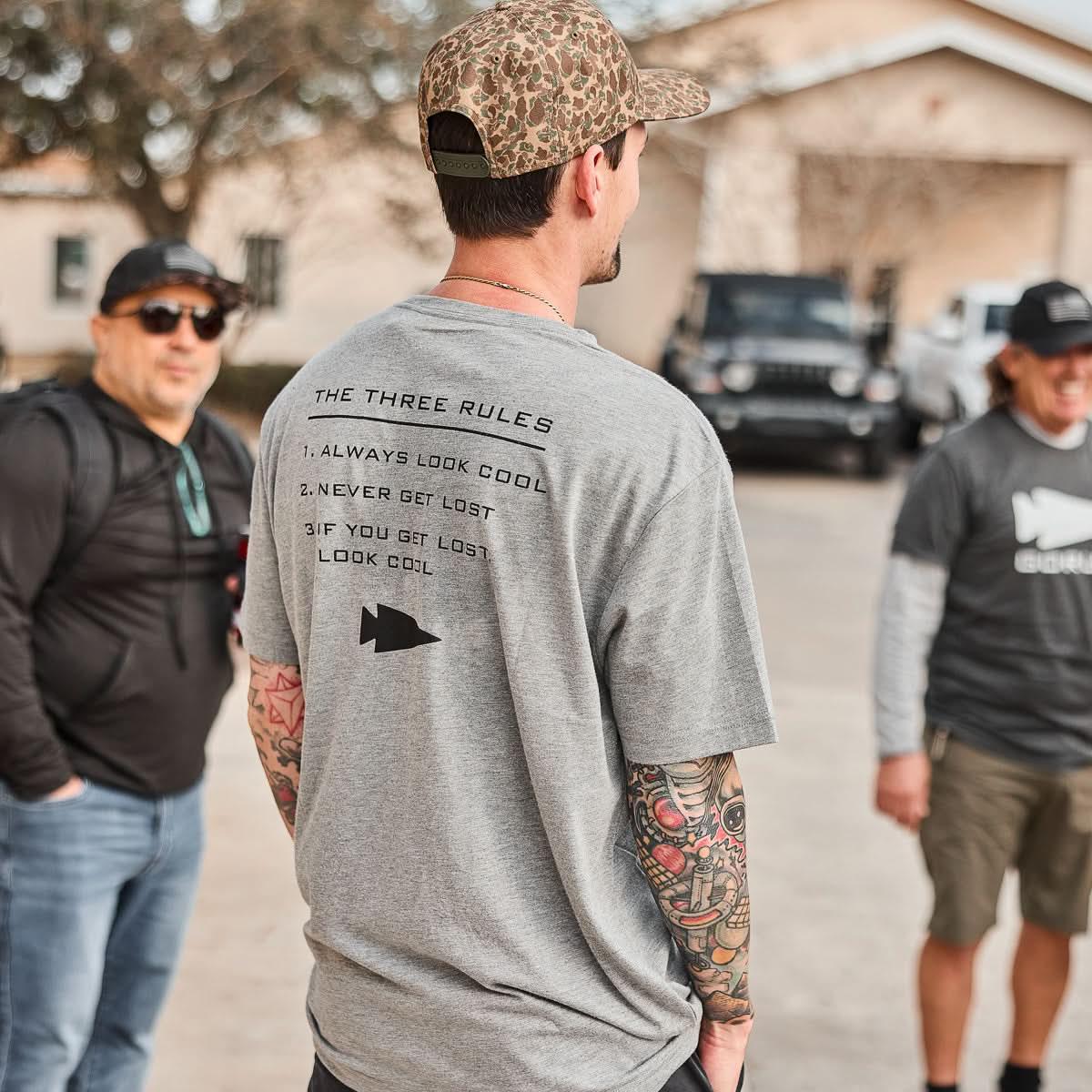 Man wearing GORUCK Three Rules grey tee with printed text, outdoors with group, casual setting