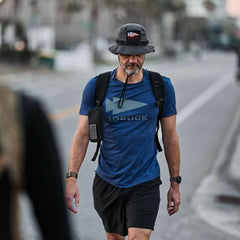 Man wearing navy GORUCK Three Rules Tee, bucket hat, and backpack rucking outdoors