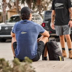 Man wearing GORUCK Three Rules navy tee sitting outdoors with rucksack, another in logo shirt.