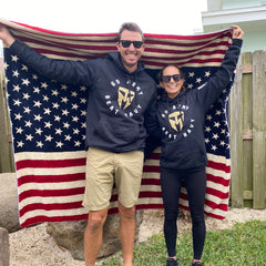 Two people wearing "Go Army Beat Navy" hoodies and Men’s Simple Shorts - Lightweight ToughDry® hold an American flag blanket behind them, smiling outdoors.