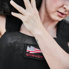 A person wearing the GORUCK Training Weight Vest 2.0, adorned with the American flag and the GORUCK logo, carries a black sandbag on their shoulder. Dust clings to the vest, while a small tattoo adorns their arm, highlighting their determination.