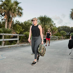 Woman rucking with GORUCK gear on outdoor path, wearing athletic apparel and carrying camo backpack