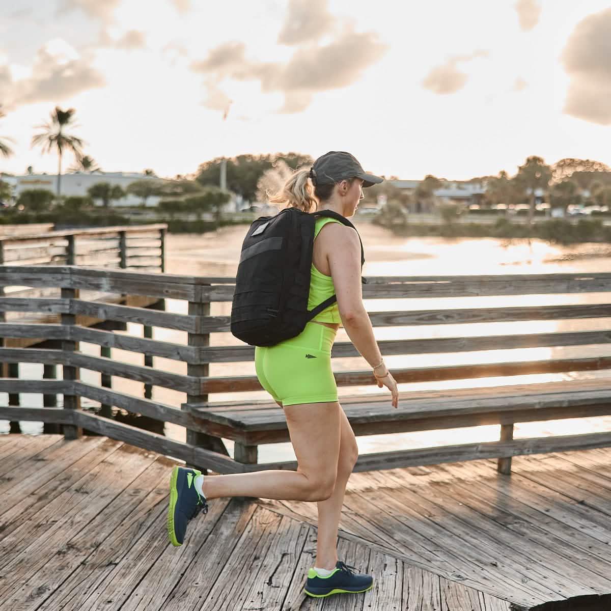 Woman in neon workout gear rucking with GORUCK backpack on boardwalk at sunset