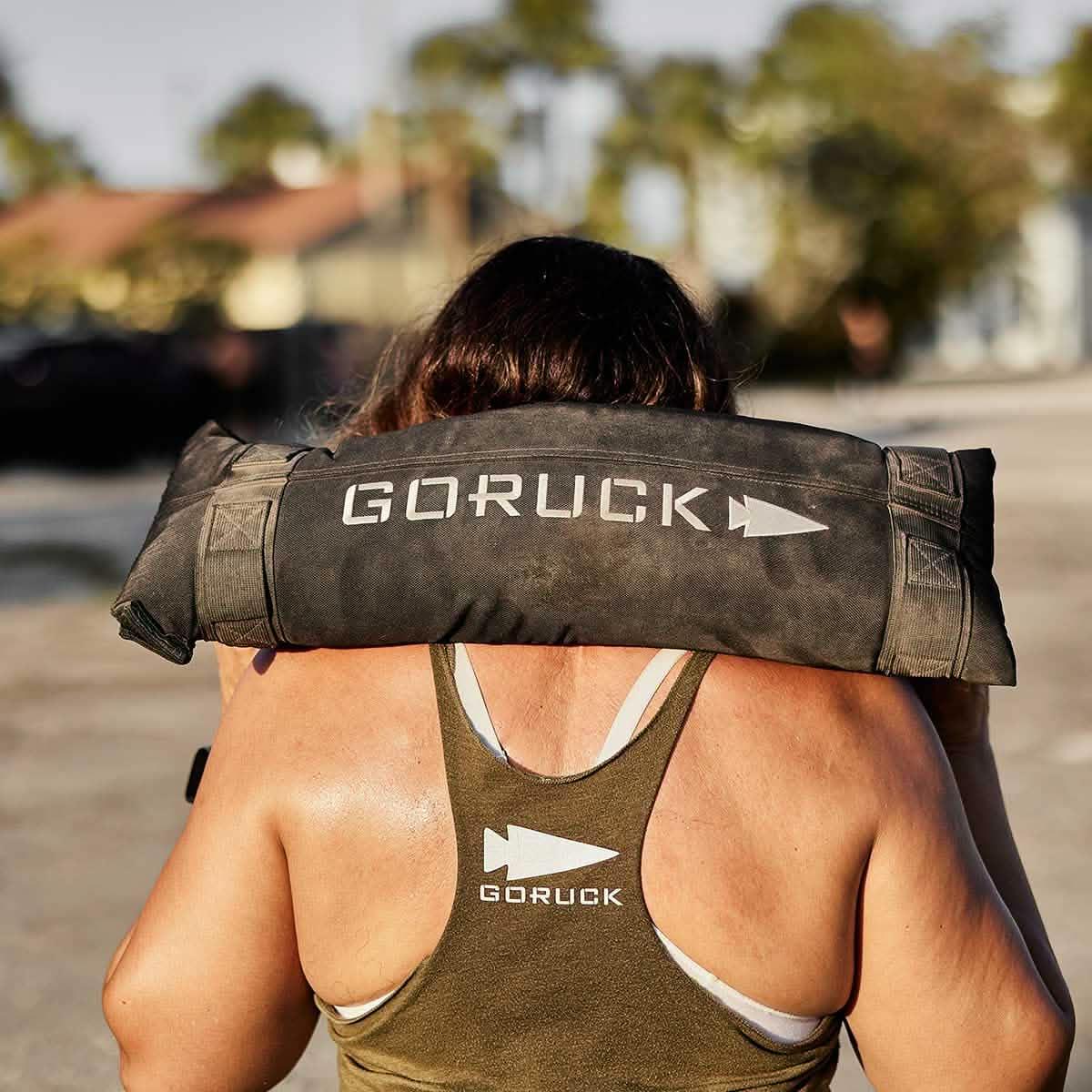 Woman carrying GORUCK sandbag outdoors, wearing GORUCK tank top, fitness training gear