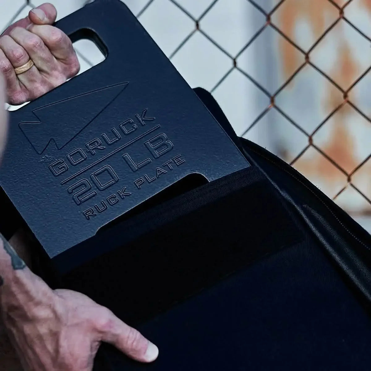 Hand placing black GORUCK 20 lb ruck plate into backpack against chain-link fence