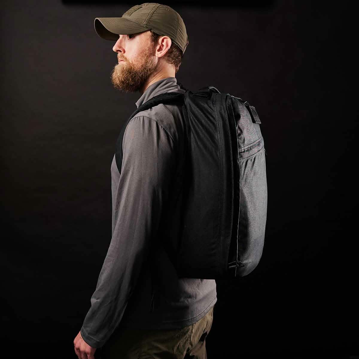 Man wearing GORUCK black rucksack, gray jacket, and green cap on black background