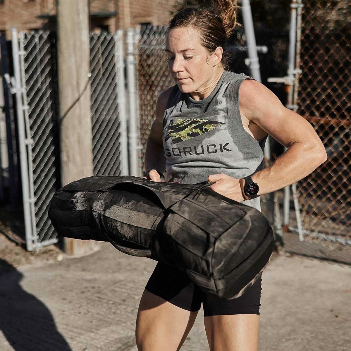 Athlete lifting GORUCK sandbag outdoors, wearing GORUCK tank and fitness watch