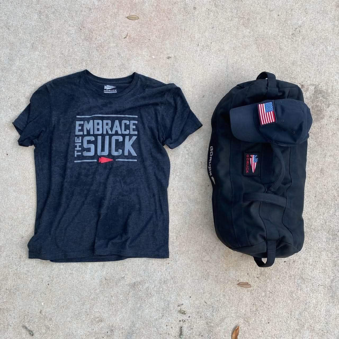 GORUCK Embrace the Suck black t-shirt, black rucksack, and cap with US flag patch on concrete