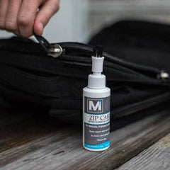Zip Care zipper cleaner bottle beside a black GORUCK bag with a hand holding the zipper