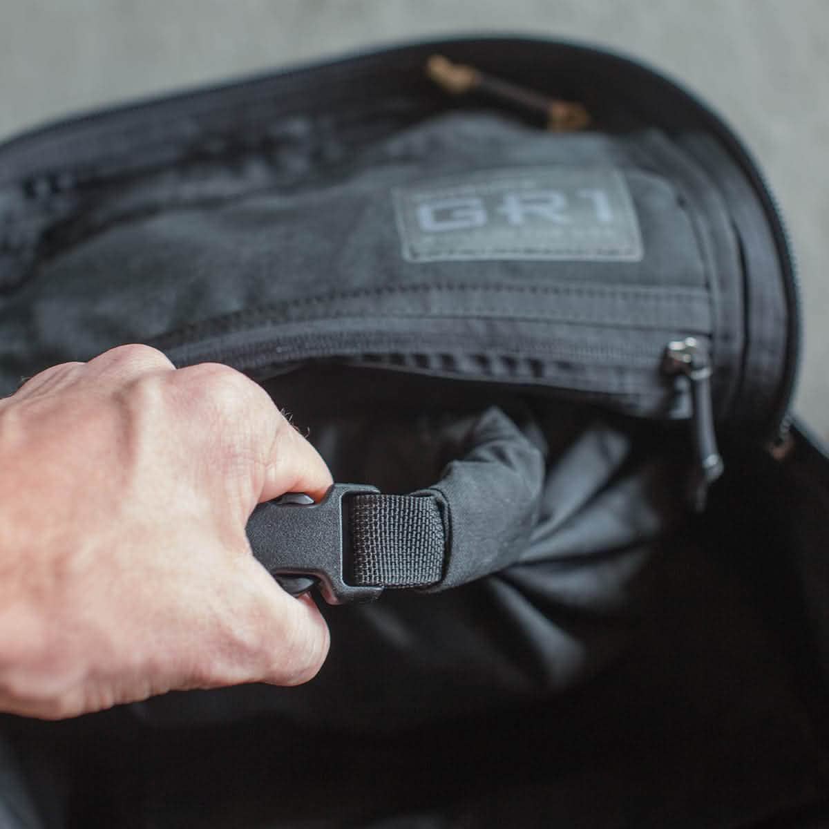 Hand holding a strap inside an open black Compression Tough Bag with a zippered pocket and visible MOLLE attachment.