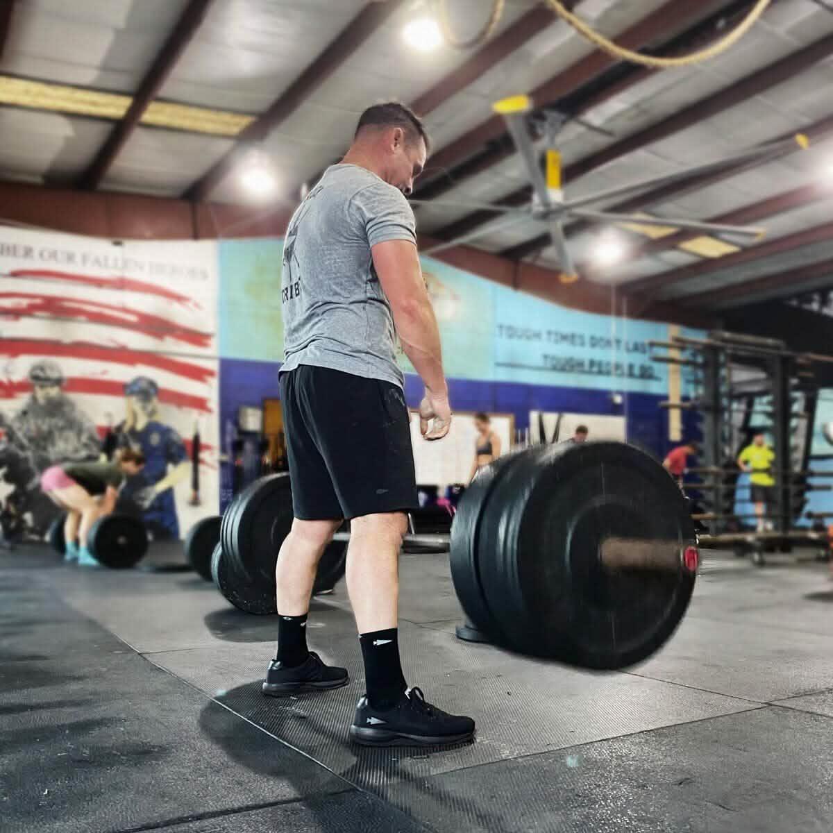 Man wearing GORUCK ballistic reflective black shoes lifting barbell in gym with mural background