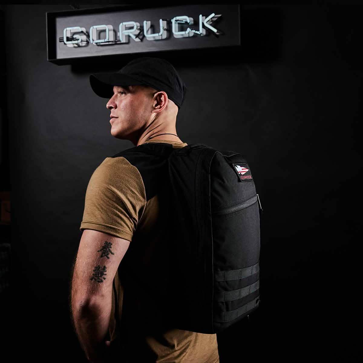 Man wearing a GORUCK black rucksack backpack, standing under GORUCK sign