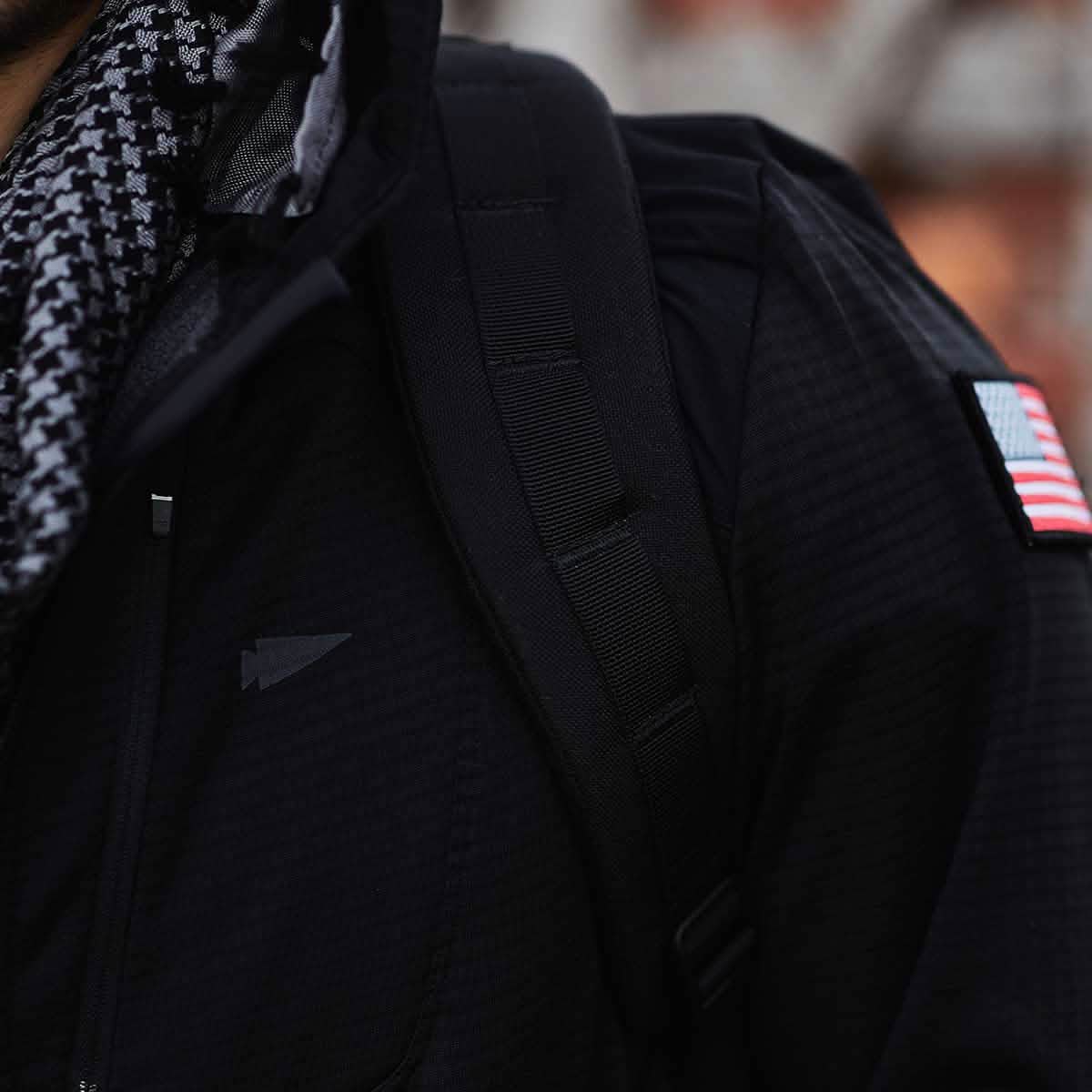 A person wearing a black GORUCK Men’s Full Zip - Polartec Grid Fleece jacket with ToughDry Fabric and a checkered scarf is visible from the chest up. They have a black backpack strap over their right shoulder, and an American flag patch adorns the sleeve.