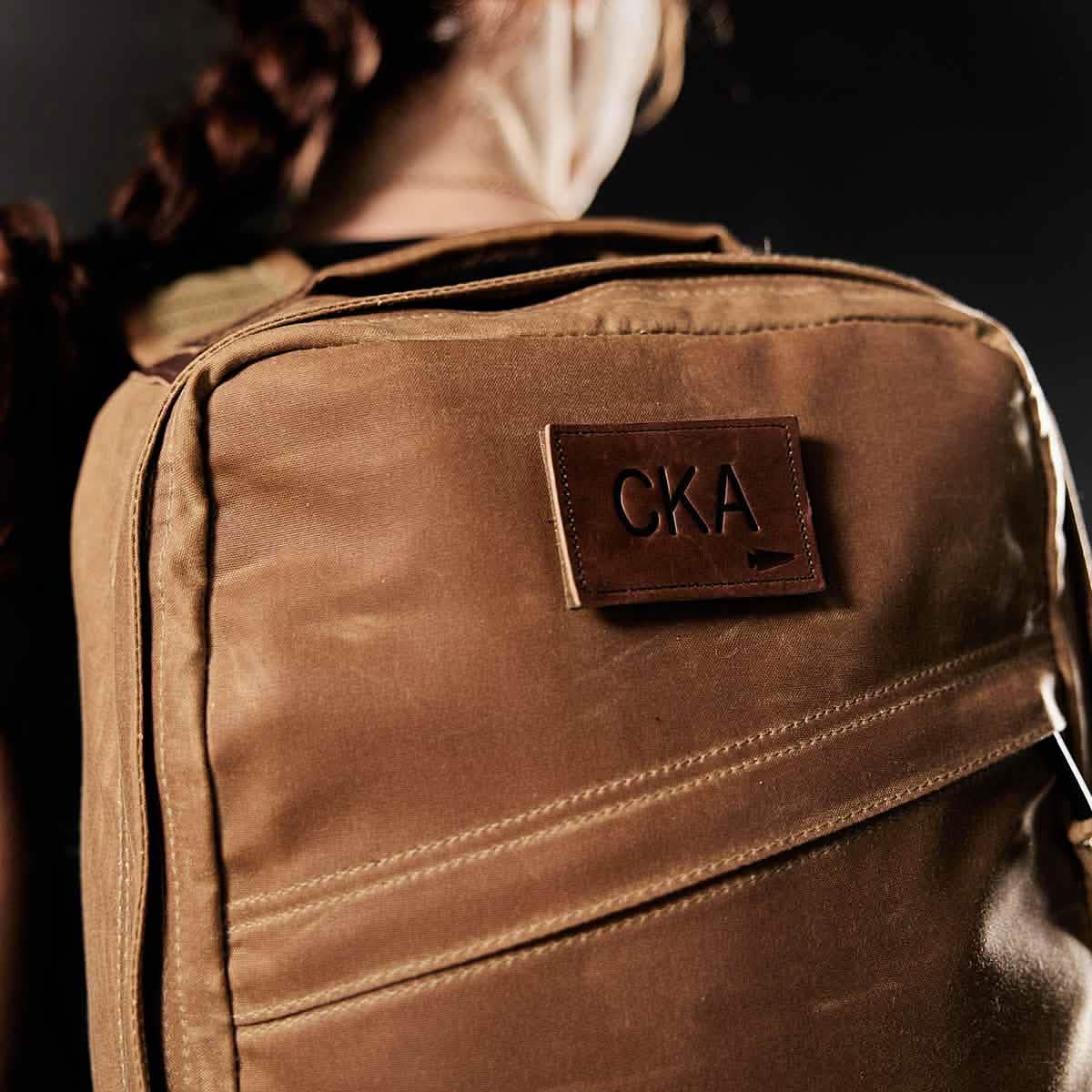 A person with braided hair is seen from behind wearing a brown backpack, which showcases a vendor-unknown Patch - Custom Leather Monogram featuring the embossed initials 