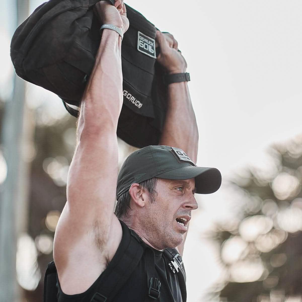 Man lifting GORUCK sandbag overhead outdoors, wearing GORUCK cap and rucking gear