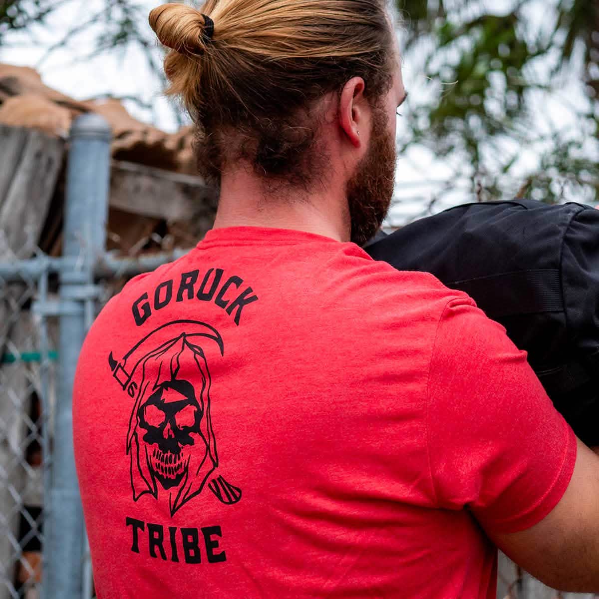 Man with long hair outdoors near a fence carries a black object while wearing the red 