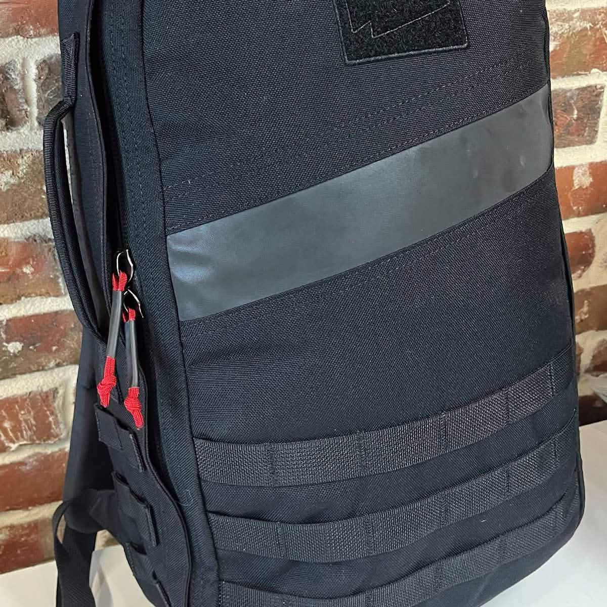 GORUCK black rucksack with reflective stripe, red zipper pulls, and MOLLE webbing, indoor setting