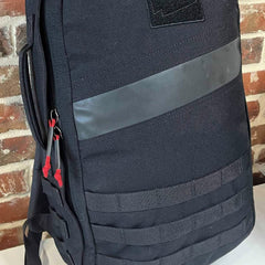 GORUCK black rucksack with reflective stripe, red zipper pulls, and MOLLE webbing, indoor setting