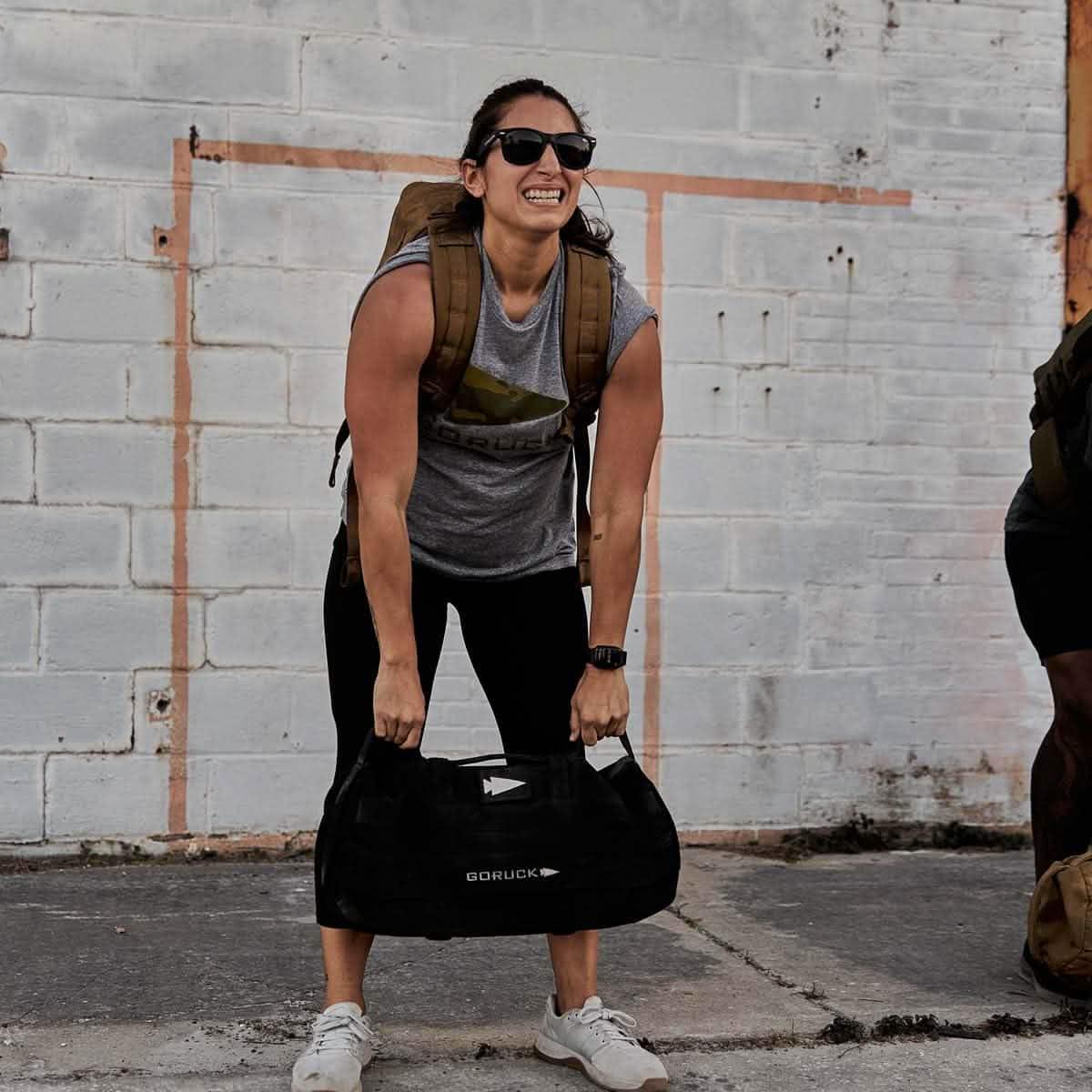 Woman lifting GORUCK sandbag outdoors, wearing backpack and workout clothing