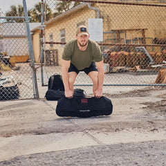 Man lifting GORUCK sandbag outdoors in front of chain-link fence, wearing green shirt and cap