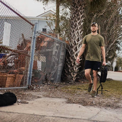 Man wearing GORUCK gear walking outdoors with rucksack, athletic shorts, and cap